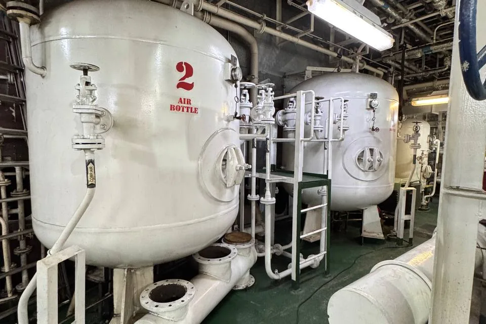 Two large vertical air receivers in a ship engine room, the nearer one stencilled AIR BOTTLE 2, with bolted manhole covers, valve stands, an access ladder and flanged pipework across the deck