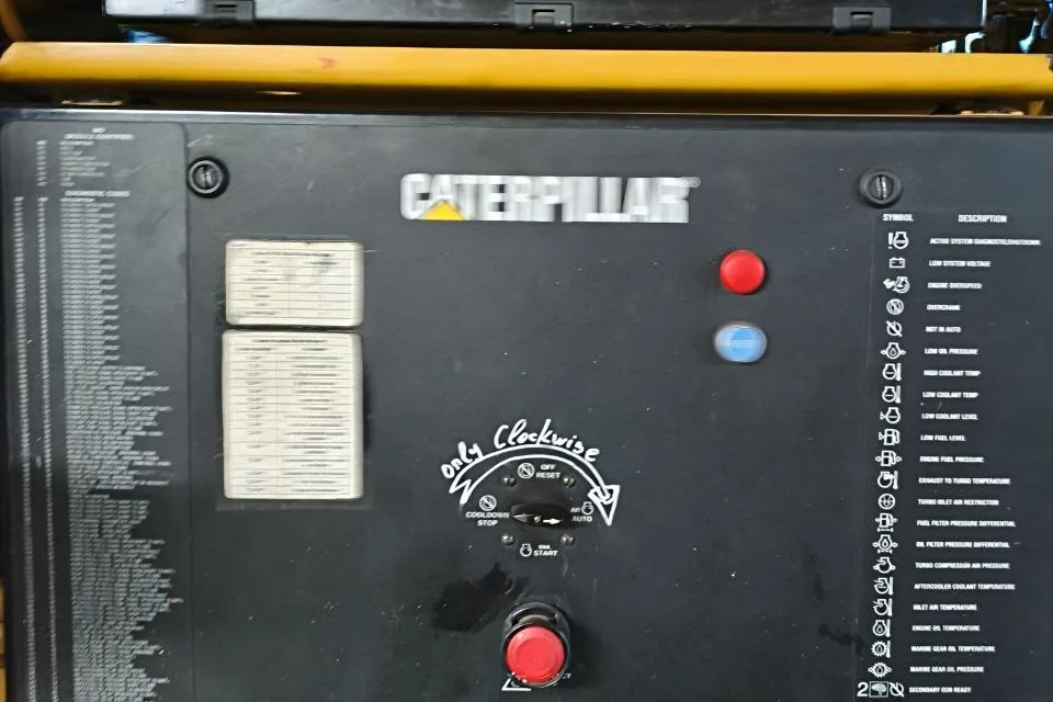 Caterpillar engine control panel aboard a vessel, with instrument gauges, an emergency stop, a handwritten note beside the key switch and a printed fault-symbol legend