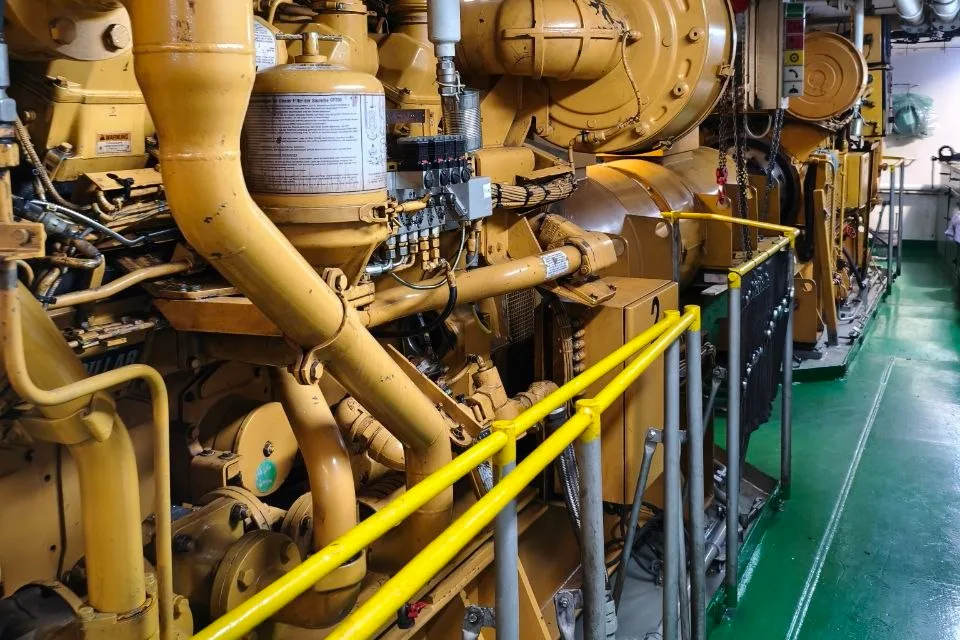 Row of Caterpillar marine auxiliary engines along a ship engine room walkway, with handrails, exhaust trunking overhead and a painted deck between the seatings