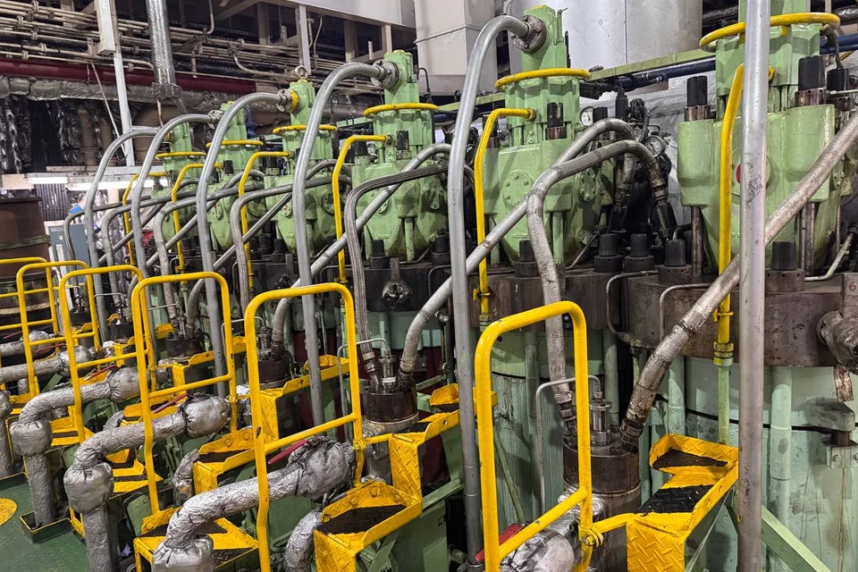 Row of cylinder units along a large two-stroke main engine in a ship engine room, with fuel pumps, lubricator lines, yellow handrails and the exhaust trunk overhead