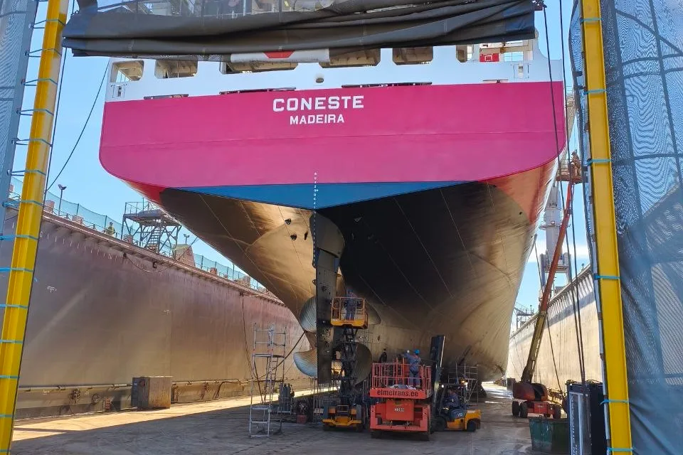 Bow, bulbous bow, propeller and rudder of a vessel seen from the dock gate while it sits in a dry dock, with elevating work platforms alongside