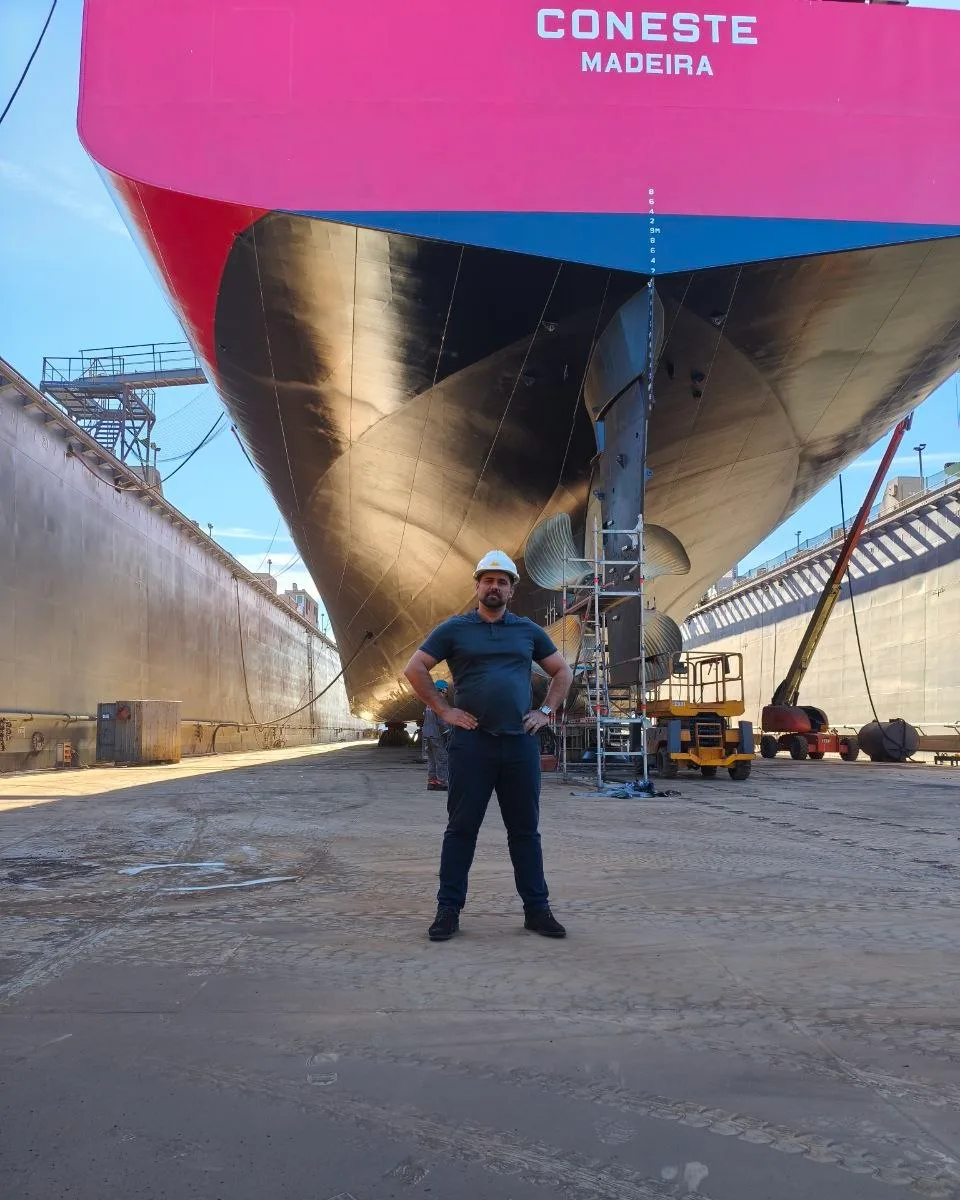 Bulbous bow and forward hull of a cargo vessel in a dry dock, with the dock walls on either side and a person in a hard hat standing on the dock floor beneath it