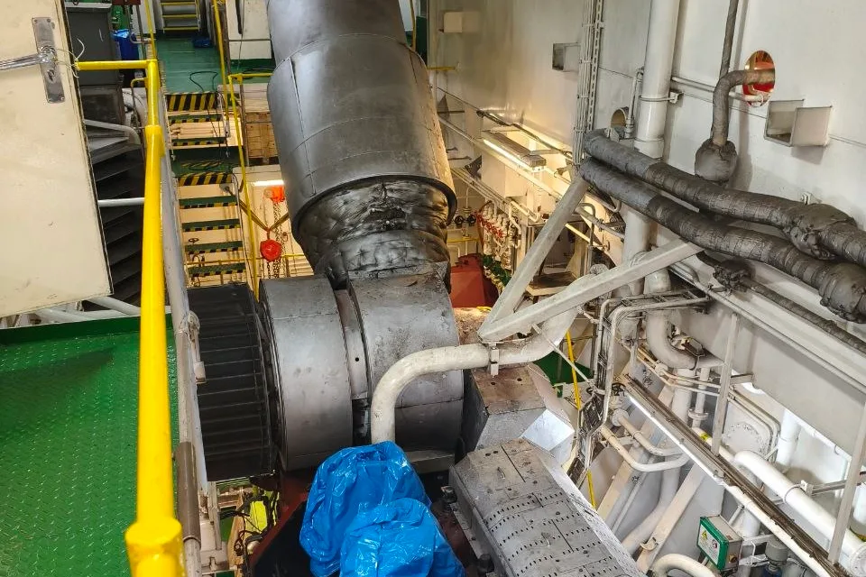 Ship engine room during an overhaul, with the main engine cylinder heads covered in protective sheeting and the turbocharger and exhaust trunk exposed above