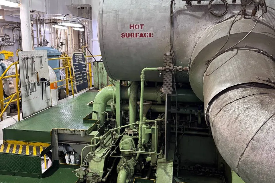 A large lagged vessel stencilled HOT SURFACE in a ship engine room, with heavy insulated ducting running from it and machinery on a green bedplate below
