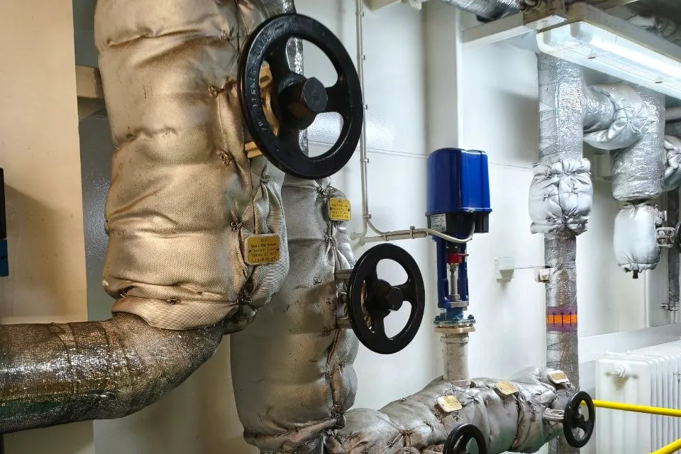 Lagged pipework running through a ship engine room with cast handwheels on two valves in the line and an actuated valve on a branch behind