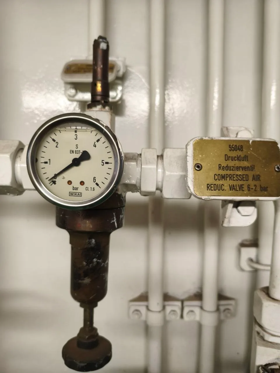 Brass equipment nameplate reading COMPRESSED AIR REDUC. VALVE 6-2 bar beside a WIKA pressure gauge on a ship's compressed air line
