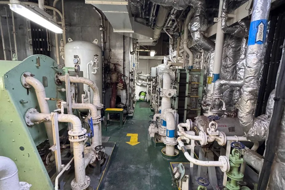 A machinery space passage with white flanged pipework, valves and a vertical vessel along one side, green painted deck and overhead cable trays