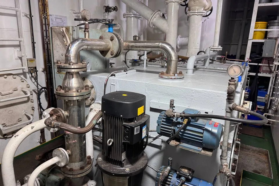 Pump group on a tank top in a ship engine room: a vertical multistage pump with its motor in front, two horizontal motor-driven pumps on a common bedplate behind, stainless suction and discharge pipework and a pressure gauge