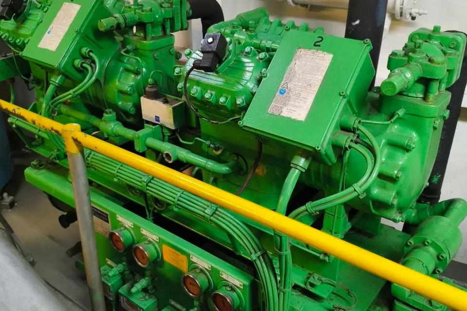 Two reciprocating refrigeration compressors numbered 1 and 2 on a common frame in a ship engine room, with terminal boxes above and a board of pressure gauges below