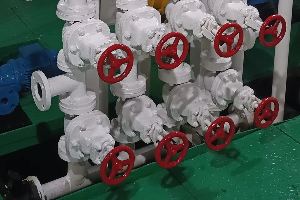Bank of gate valves with red handwheels on white flanged pipework in a ship engine room, pump alongside on green painted floor plates