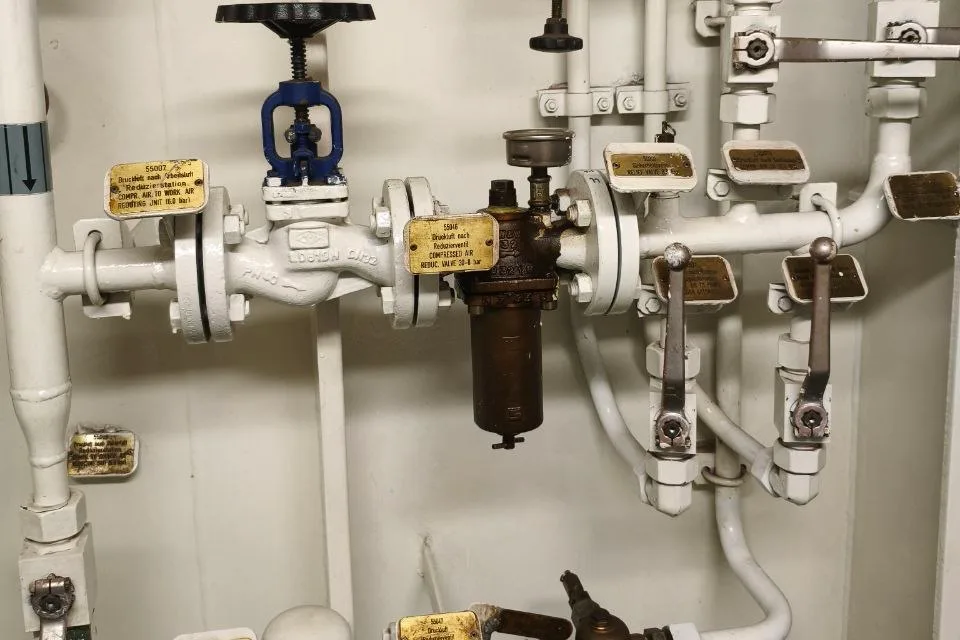 Bulkhead-mounted valve and strainer station on flanged white pipework in a ship engine room, with a gate valve, a bronze strainer bowl and engraved line tags on brackets